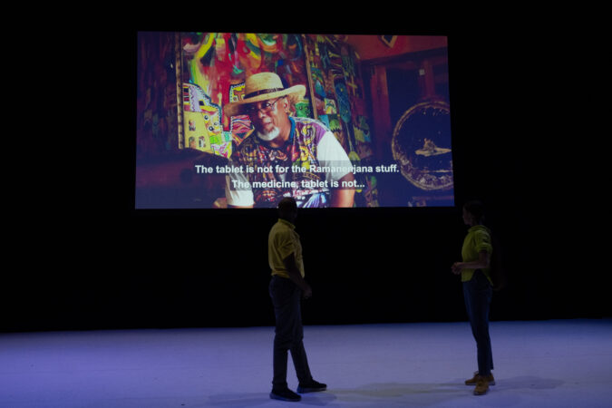 Two performers stand silhouetted onstage. Projected behind them is a person sitting a brightly patterned room, captioned “The tablet is not for the Ramanenjana stuff The medicine, tablet is not…”