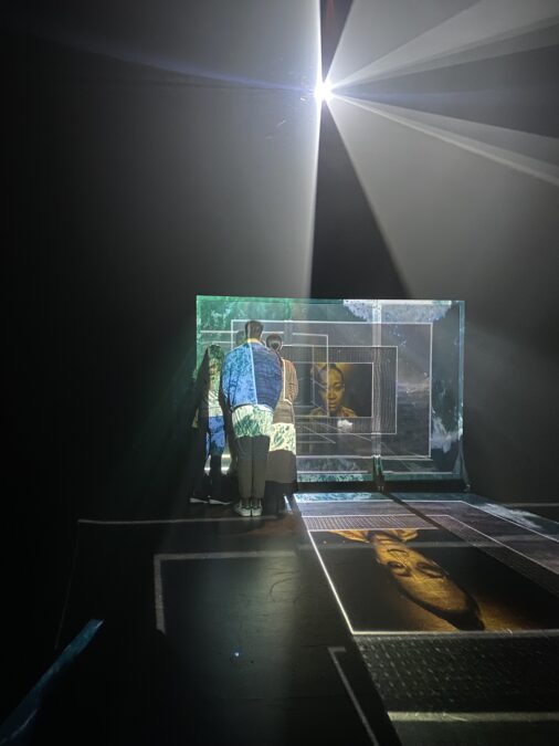 Three people stand observing a transparent screen projected with images that reflect back onto the dark floor. The projections show a face tinted yellow, outlined by several frames of atmospheric green, white, and blue textures. A spotlight cuts through the dark background.