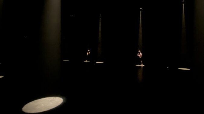 A dark stage illuminated by occasional spotlights. Under two of the spotlights stand silhouetted figures.