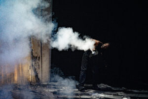 A trail of fog emits from a smoke machine, held by a Black person wearing black pants whose mouth is open in a smile or call. A ink-covered canvas tarp sprawls on the ground.