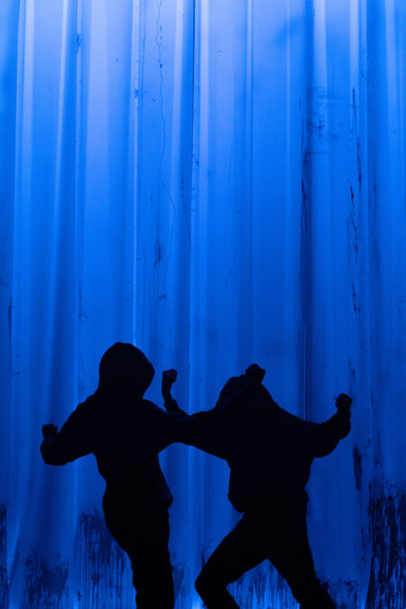Two silhouetted people dance with arms raised, against a tall blue streaky backdrop.