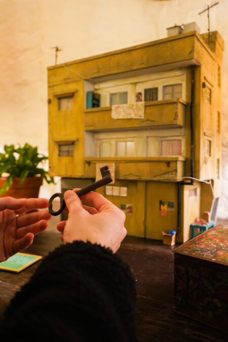 Closeup of a hand holding a large metal key. In the background on a table sits a miniature replica of an apartment building, a potted plant, and ornamental box.