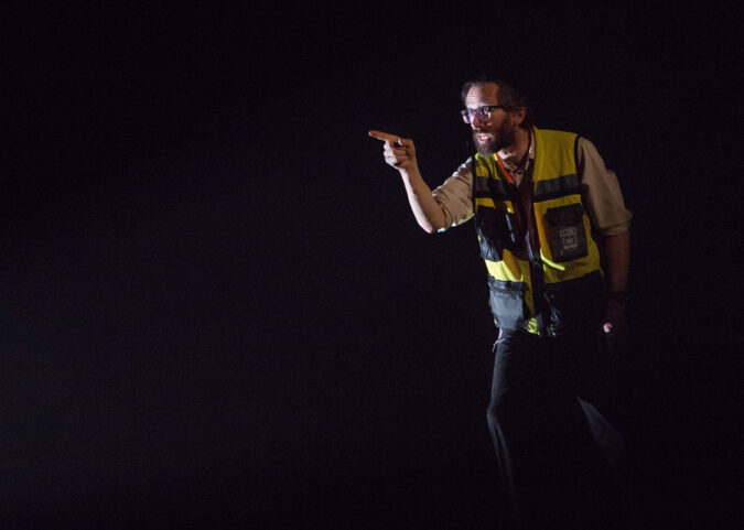 A man in a reflective safety jacket talks while pointing a raised finger at an unseen subject. The background is cast in shadow.