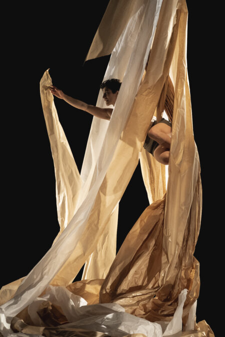 A performer hangs suspended in midair, hand outstretched upward as if flying. Draped around them are large swaths of paper that capture light shining through.