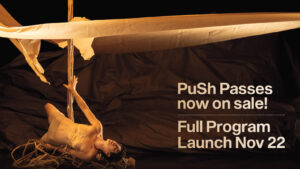 “PuSh Passes now on sale! Full Program Launch Nov 22”. A performer lies on a pile of twisted branches holding up a wooden pole struck through floating layers of paper.