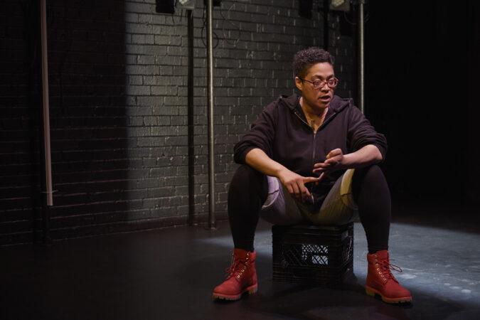 Donna-Michelle St. Bernard, a Black woman wearing red eyeglasses, sits on an upturned milk grate, counting on her fingers and speaking.