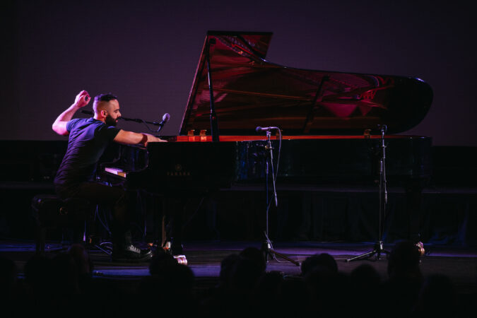 A person with short hair and beard, wearing a black shirt, plays enthusiastically on a grand piano in to a shadowed audience.