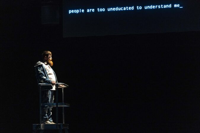 A tall, barded person stands at a raised metal platform, suspended in air. They are wearing ripped denim jacket and jeans, with a grey hoodie and headphones around their neck, illuminated by a spotlight. A screen behind them reads “people are too uneducated to understand me_”