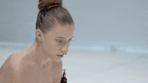 Closeup of a naked person with brown hair in a tight bun, that cuts to them crouched and leaning over with arms outstretched to reveal an electronic device on their arm.