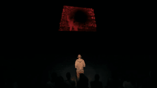 A person stands onstage facing a projected screen, the silhouettes of a seated audience behind them. On the screen grainy red video footage plays.