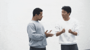 Jean Abreu and Naishi Wang, both dressed in dirty sweatshirts, converse rhythmically and enthusiastically, making emphatic hand gestures towards each other.