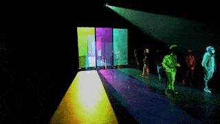 A triptych of transparent screens with magenta, turquoise, and yellow projections casts long reflections on the ground. Observers walk around the dark room, bathed in the light of the projections.