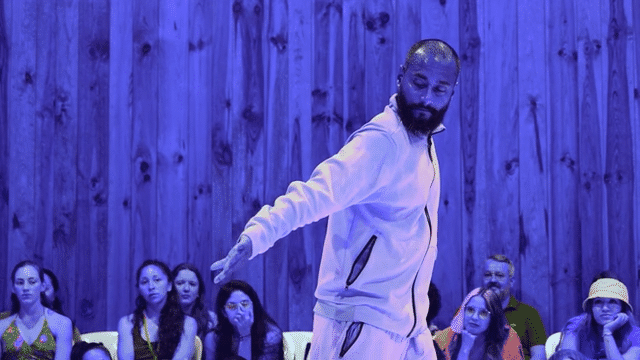 Rakesh Sukesh, with shaved head and beard, twists his torso while facing backward, arm outstretched. He is wearing a tracksuit and the room is bathed in purple light. In the background, several people sitting against a wood wall watch raptly. Subtitled “She said why don’t you make this more sophisticated”