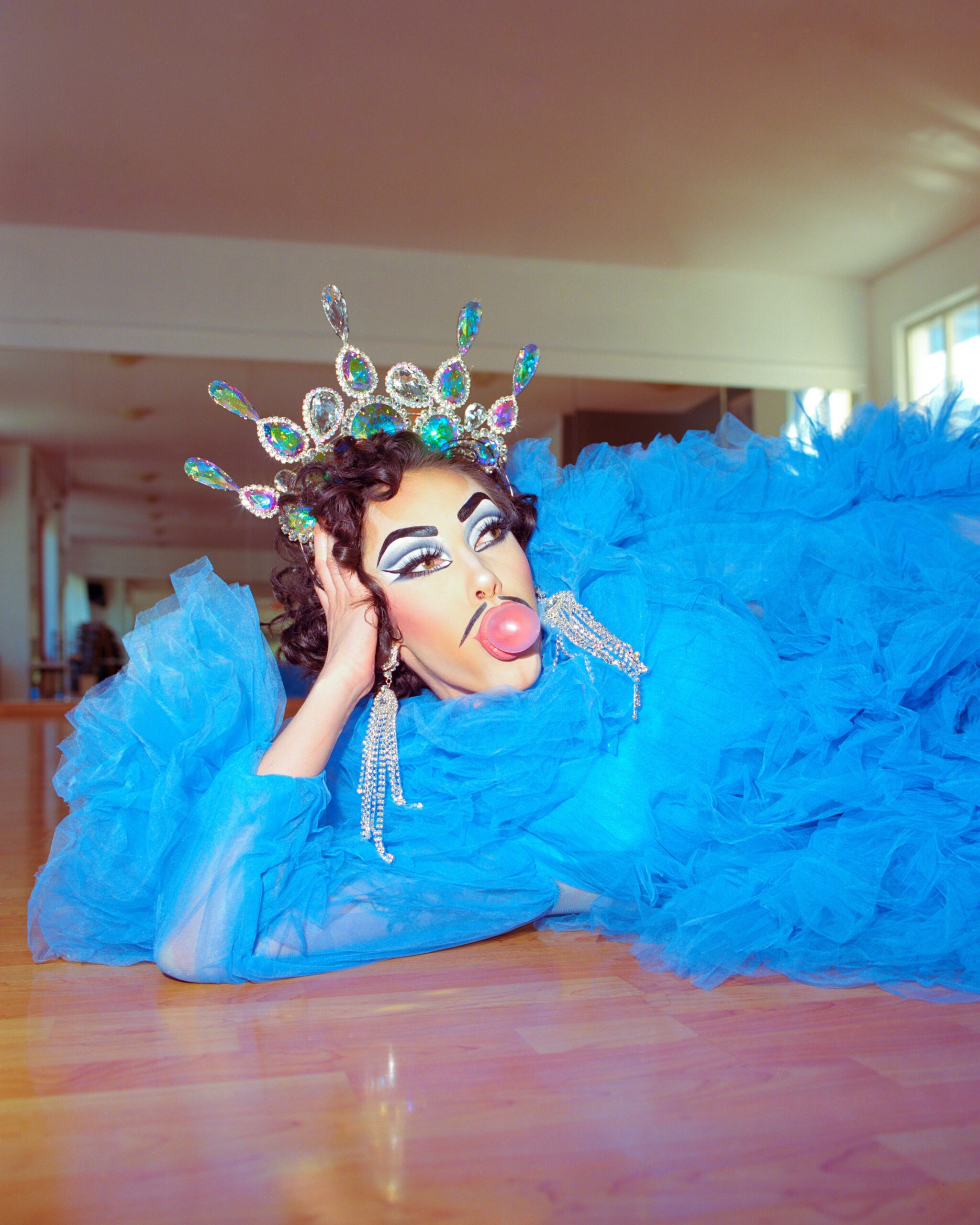 Genesis lying on their side on the floor wearing a blue dress, a tiara with blue stones, and blowing a gum bubble.