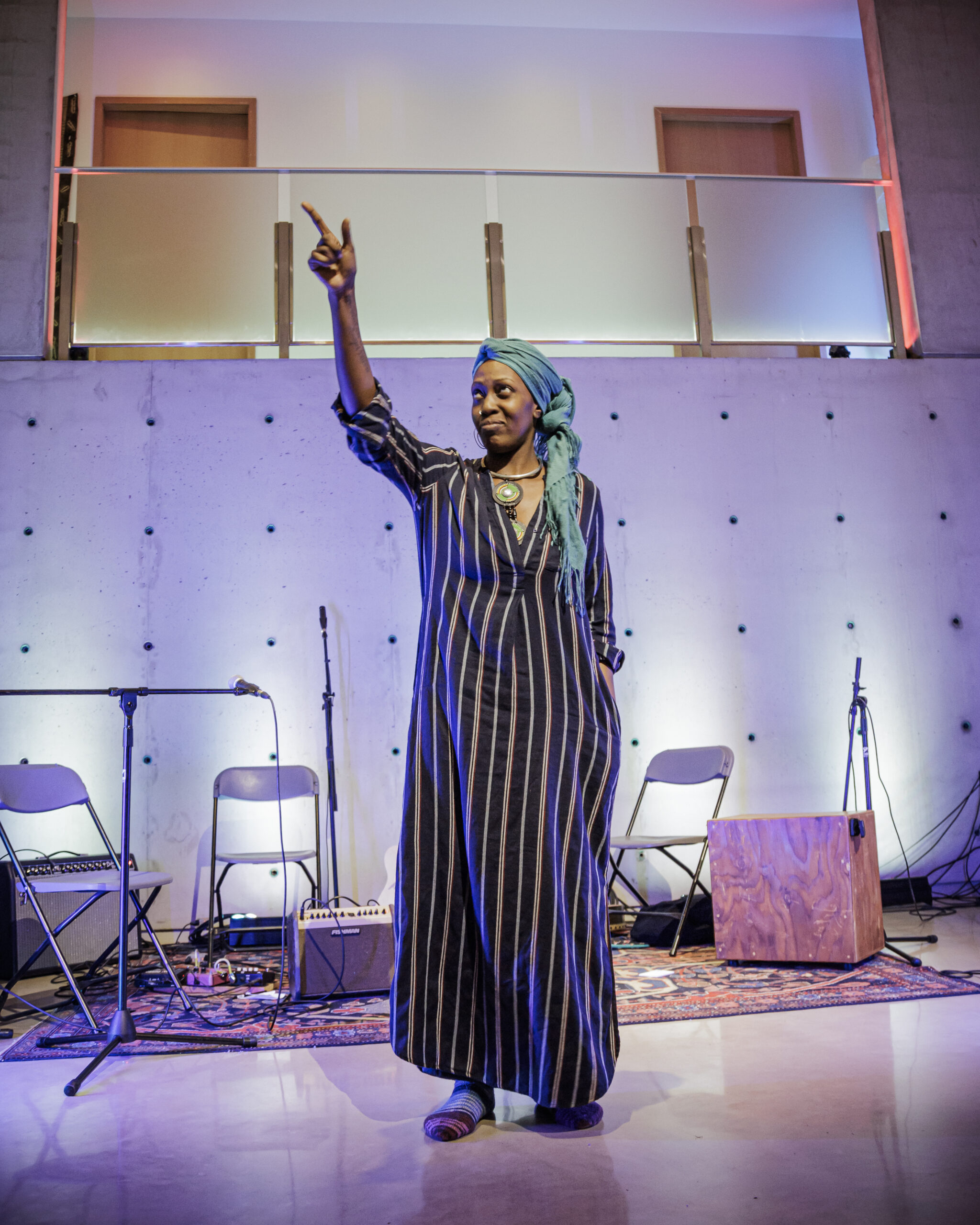 Tony standing on a stage with a few chairs and sound gear behind them, wearing a long striped dress and turquoise headscarf.