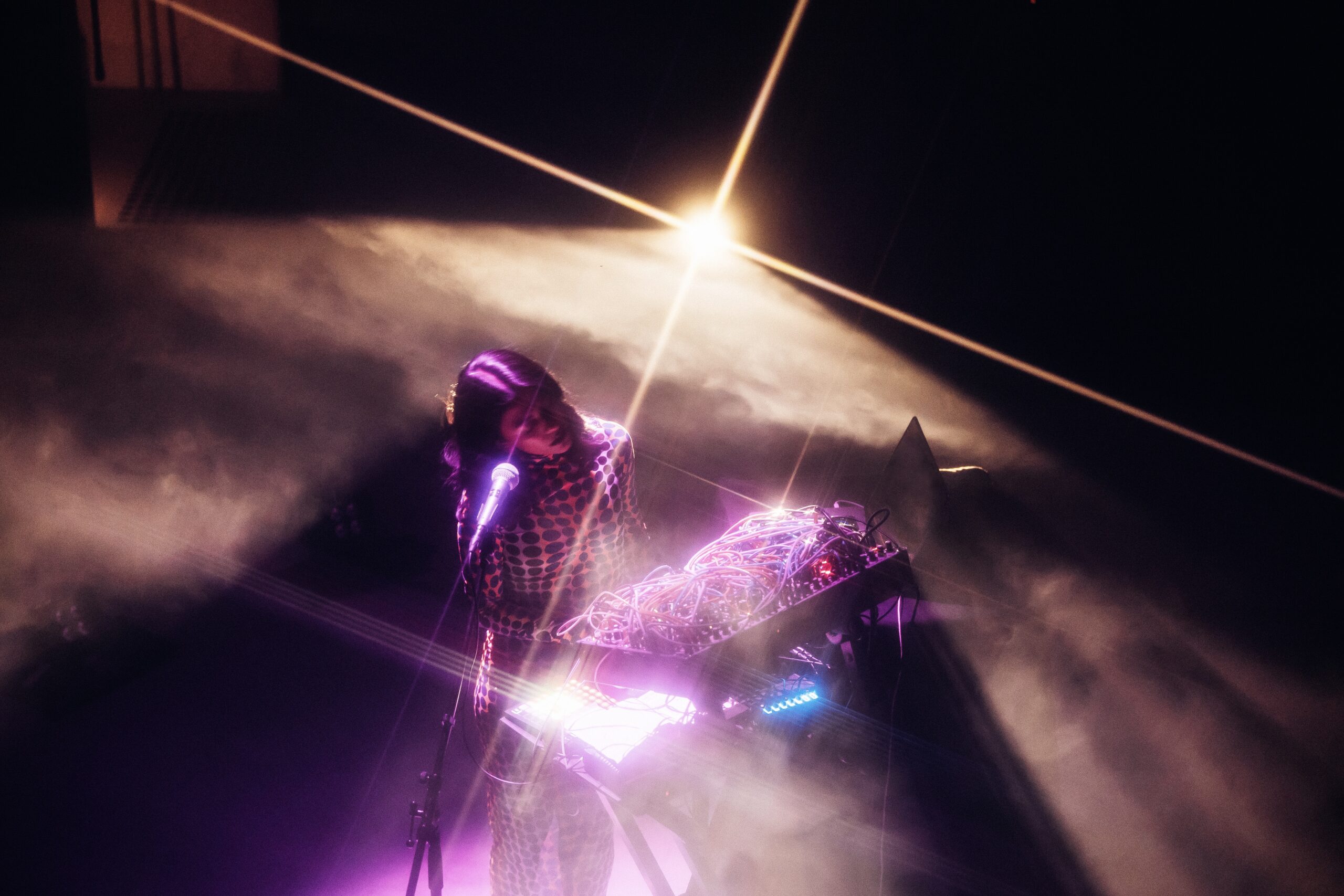 Overhead shot of a woman performing with a microphone and console with a complex array of wires, with creative spotlighting and a  background starlight flare.