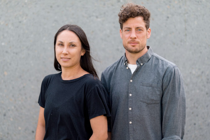 Portrait of Lisa Mariko Gelley wearing a black shirt and standing in front of Josh martin wearing a grey button down, standing against a grey wall