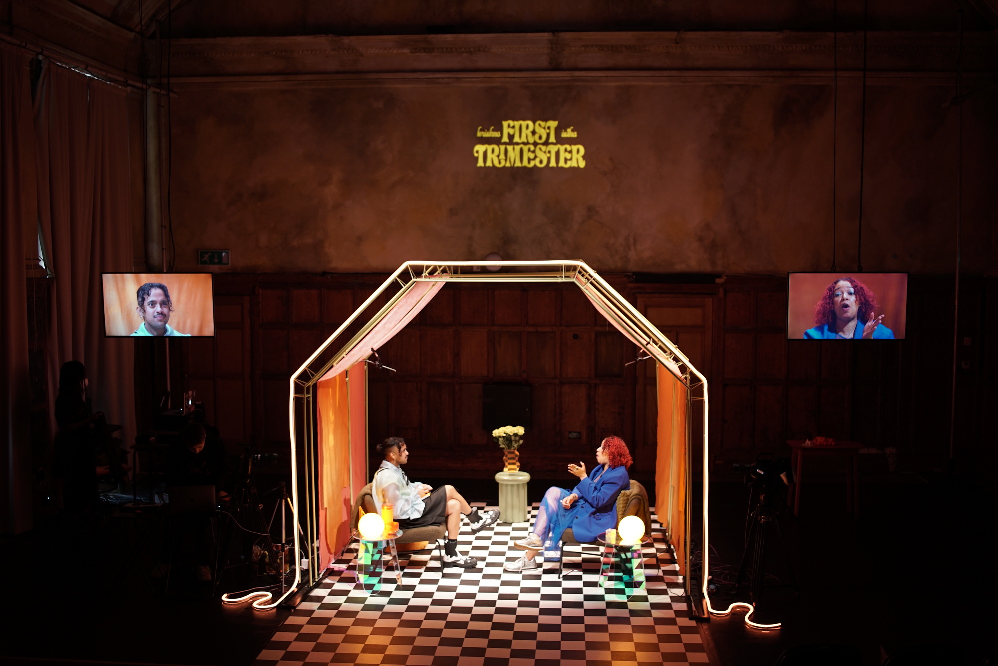 Wide shot of two performers sitting across from each other on a stage in a living-room set. The left figure is wearing a white bouse and black bottoms and is loooking at the performer on the right, who is wearing a blue suit, has red hair and is speaking. The set has a checkered floor, neon lights outlining walls and a ceiling, and video monitors of the performers. In the background "First Trimester" is projected onto the wall.