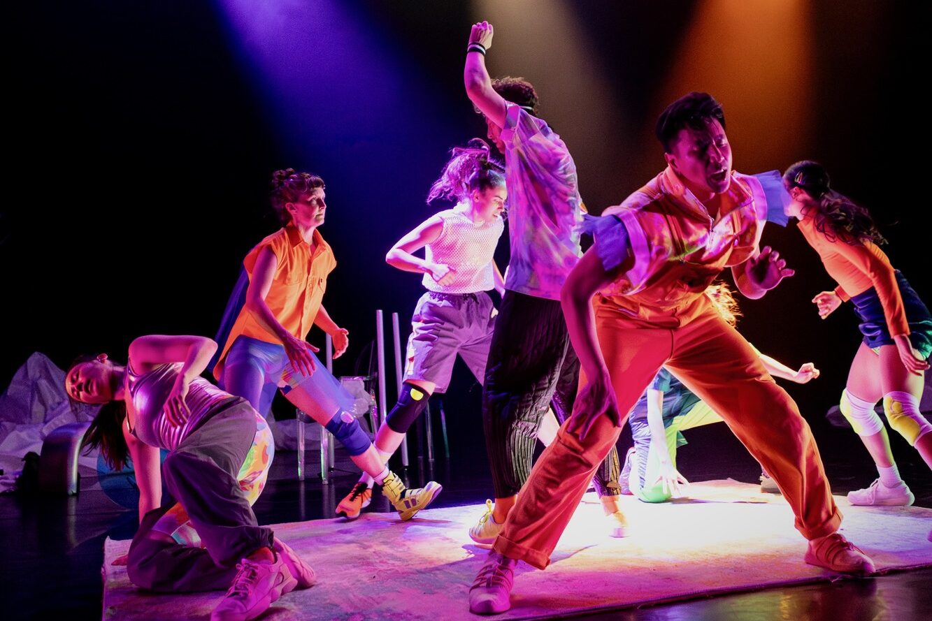 Performers dancing on a stage, lit by pink and purple light