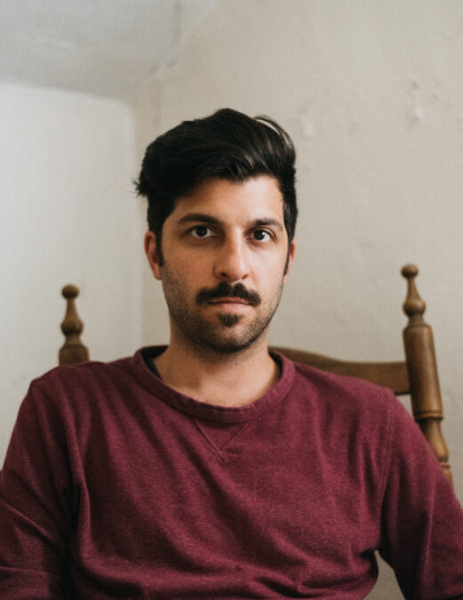 Portrait of Adam Paolozza, who has dark hair, a moustache and beard, and is wearing a burgundy shirt. 