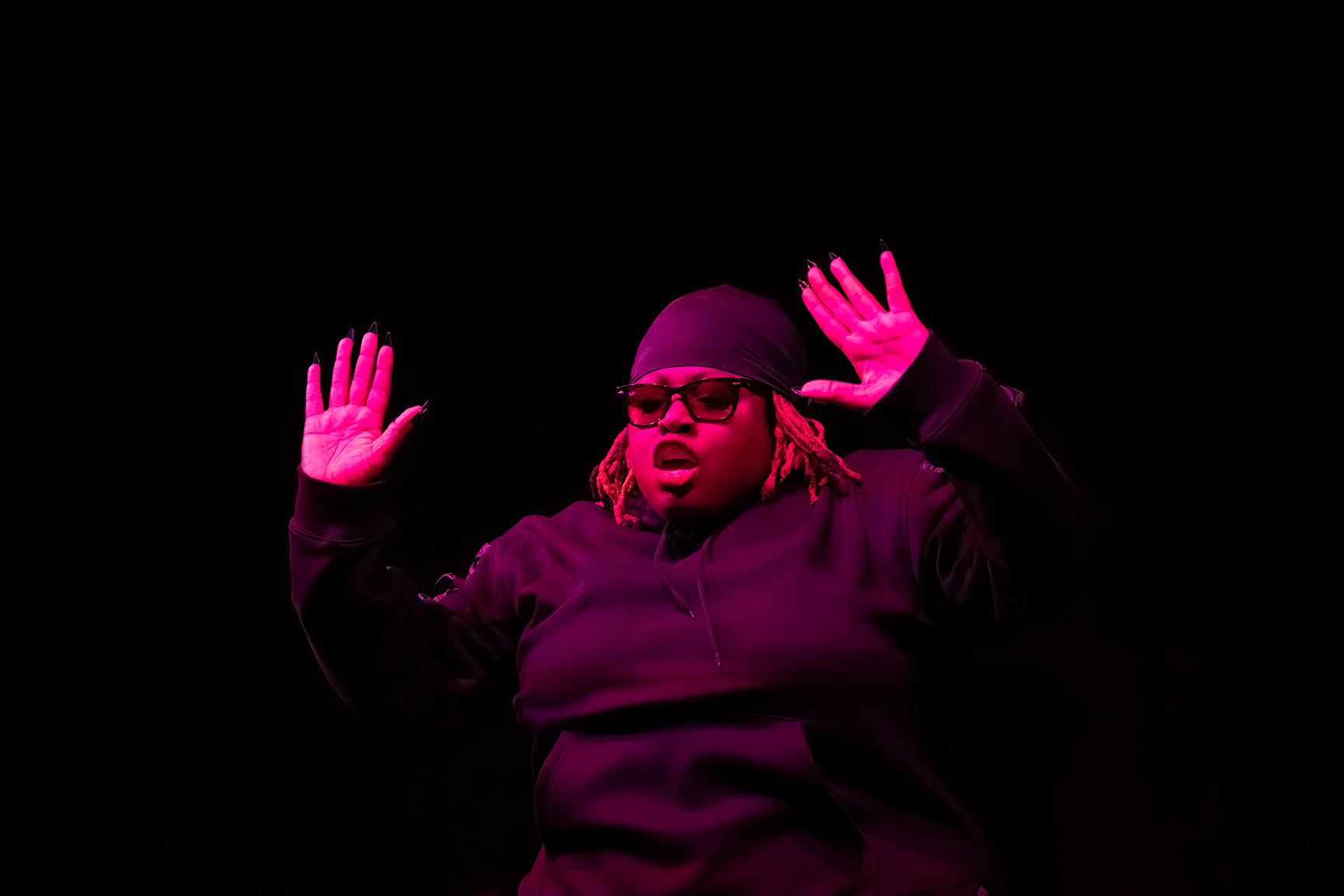 Alexandra 'Spicey' Landé, wearing a durag and black glasses, lit in pink light with hands raised.