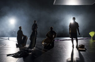 The silhouettes of four dancers break up a dark stage strewn with long swaths of fabric and geometric sculptural shapes. They stand and kneel, each holding different objects, like a box, a long piece of cloth, and a plank of wood dragged by a wire handle.