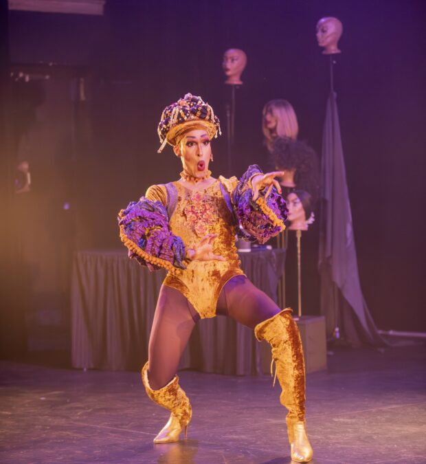 A south-asian drag artist wears a gold, crushed-velvet leotard with matching knee-high high-heeled boots. The artist is caught mid gesture and mid word, reaching out with a look of shock on their face. In the background, an array of wigs perch atop a small cluster of mannequin heads.