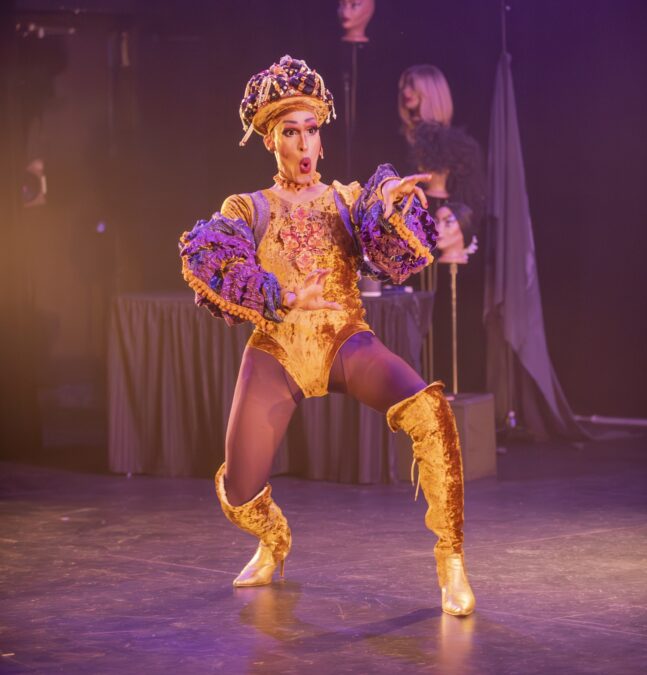 A south-asian drag artist wears a gold, crushed-velvet leotard with matching knee-high high-heeled boots. The artist is caught mid gesture and mid word, reaching out with a look of shock on their face. In the background, an array of wigs perch atop a small cluster of mannequin heads.