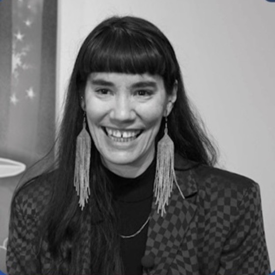 Black and white portrait of Camille Larivée. They have long dark hair with bangs, and are wearing long beaded earrings and a checkered blazer.