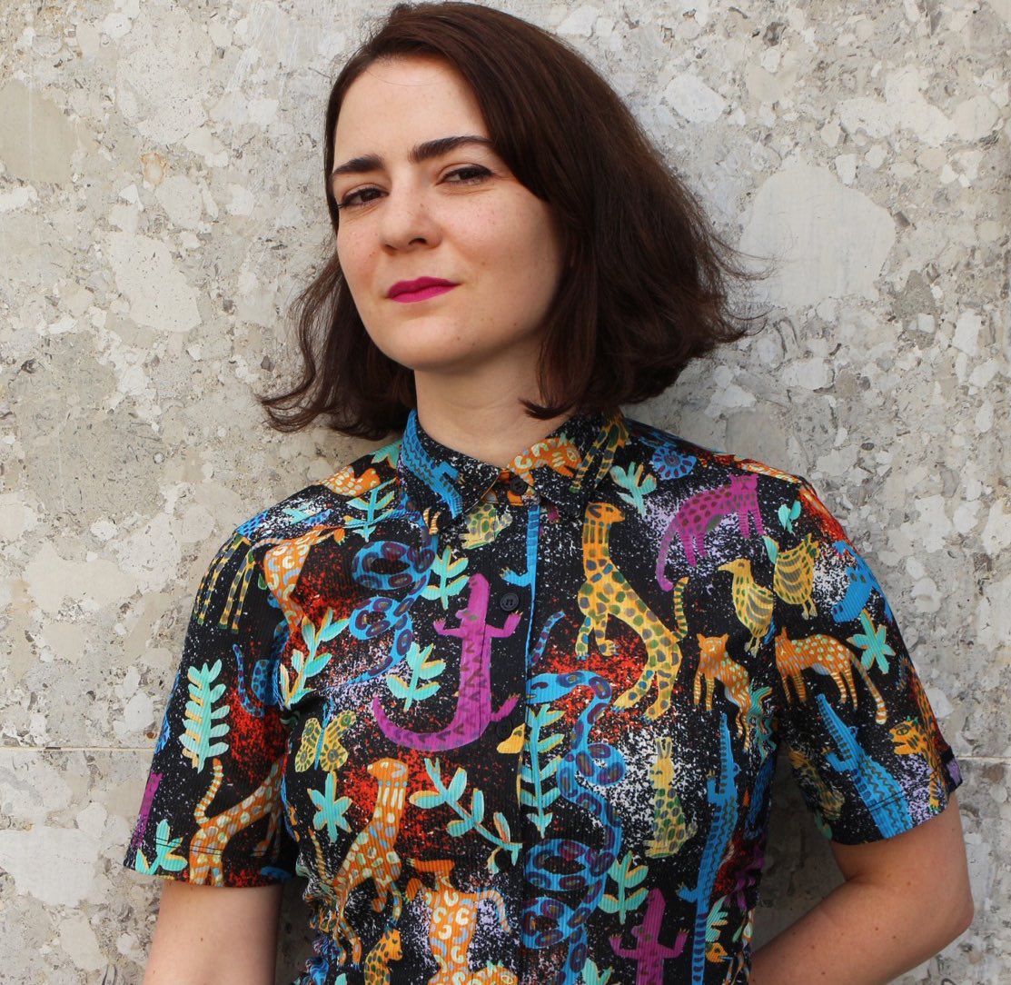 Cecilia Kuska posing against a stone wall. She has shoulder-length brown hair and is wearing red lipstick and a button-up shirt wth brightly patterned animals and plants.