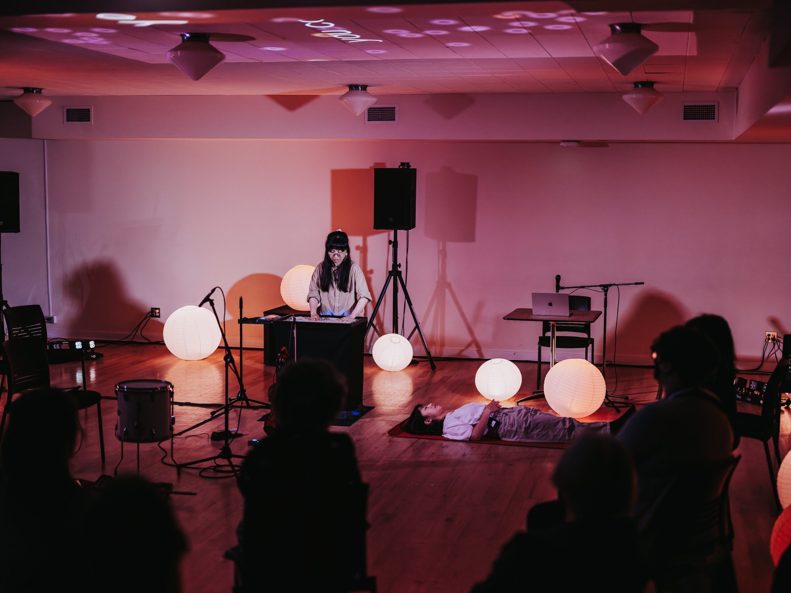 A performer with long dark hair stands onstage at a podium. On the floor is another performer lying down, sound equipment and white ball lights. Silhouettes of the audience line the bottom of the frame.