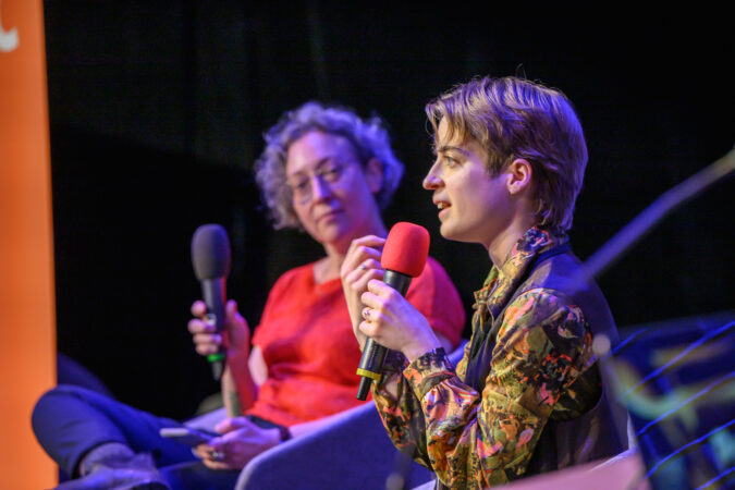 Two seated people in a dark room, bathed in purple light and holding microphones. The person on the left is out of focus and has short grey hair, glasses and a red shirt, looking at the speaker. The person on the right has short blonde hair and is wearing a green and brown patterned blouse, and is speaking into the microphone.