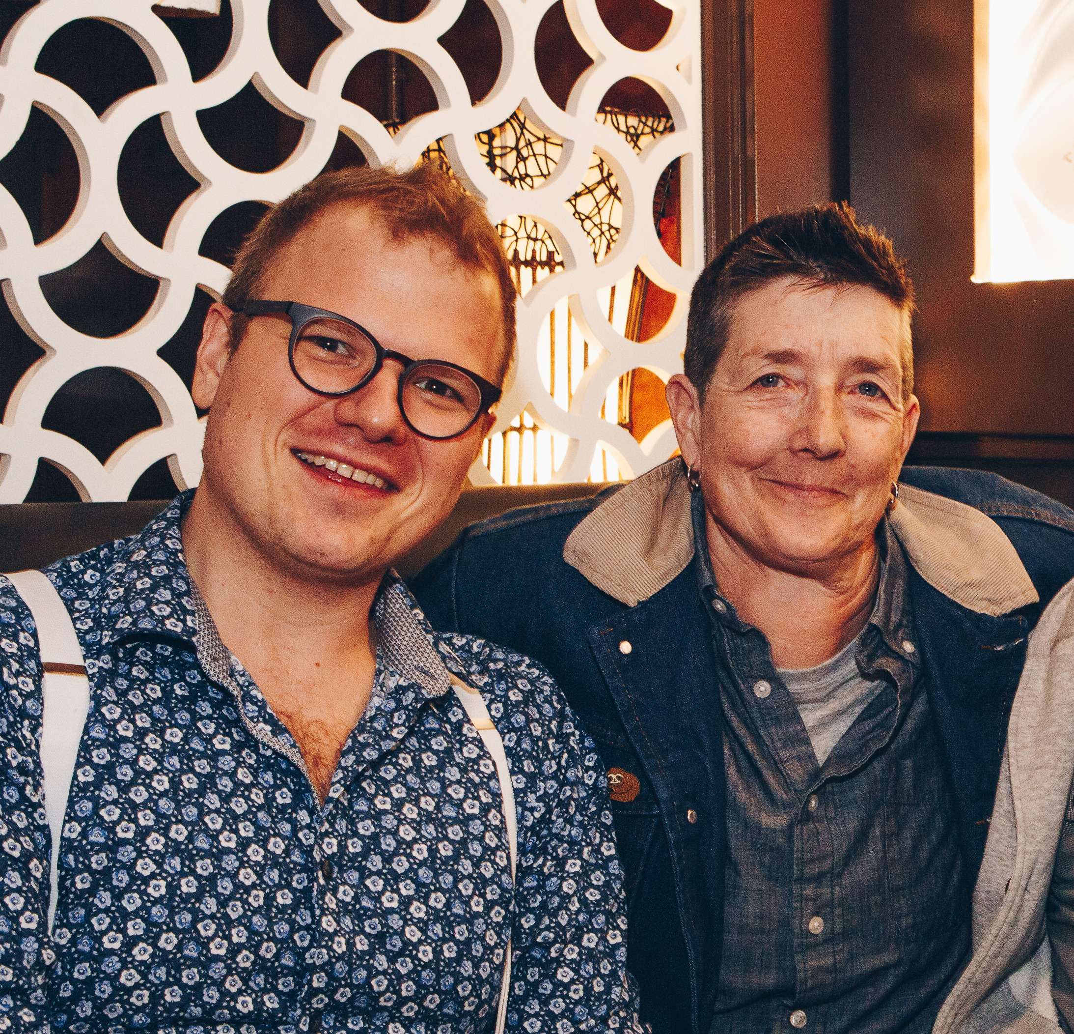 Dave Deveau and JD Derbyshire together smiling at the camera. On the left, Dave wears black glasses, blue floral shirt and white suspenders. JD wears a denim jacket and gray button down shirt.