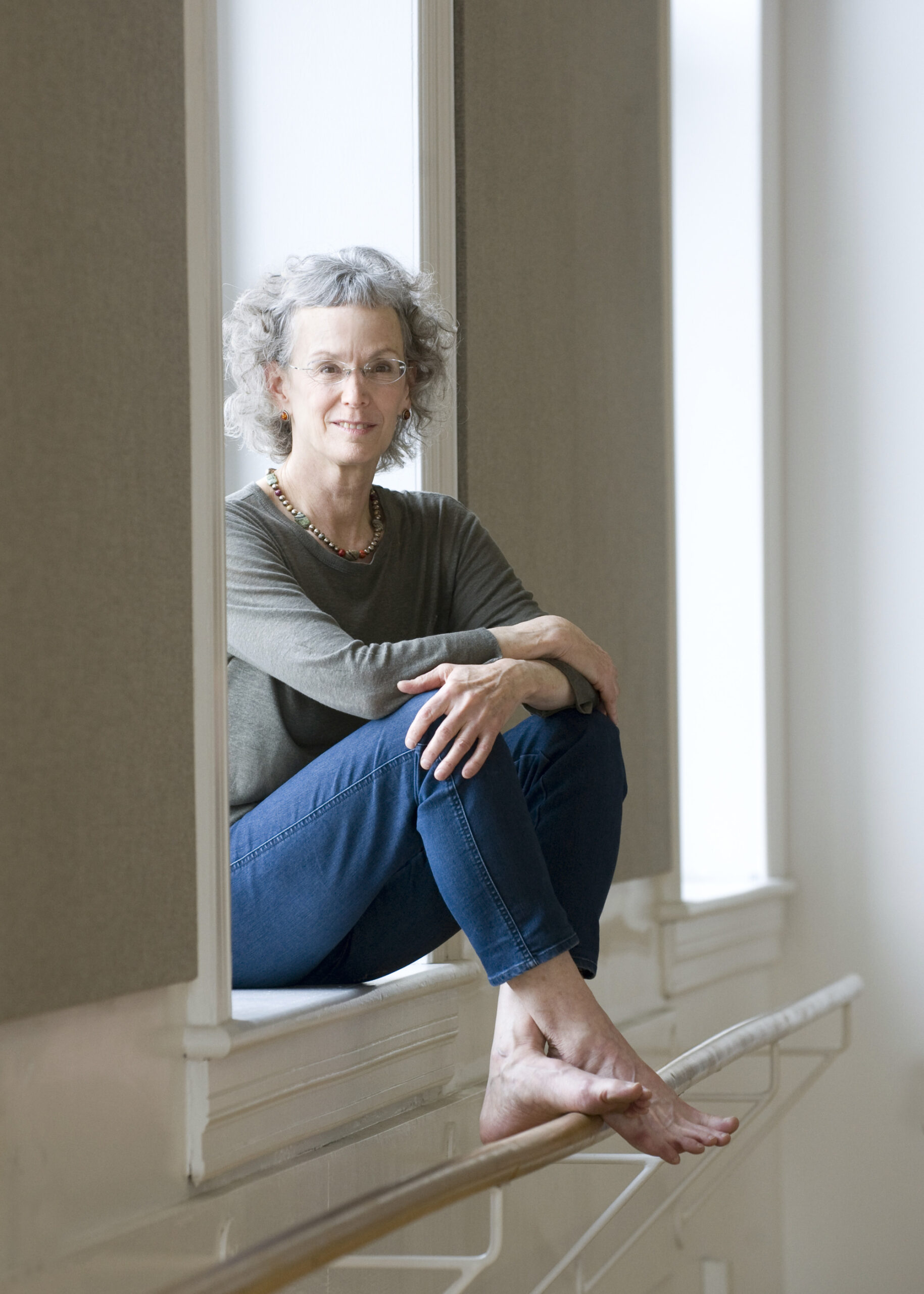 Dena Davida sitting in a window ledge, bare feet touching a ballet barre. She has curly gray hair and is wearing a grey t-shirt, beaded necklace and blue jeans.