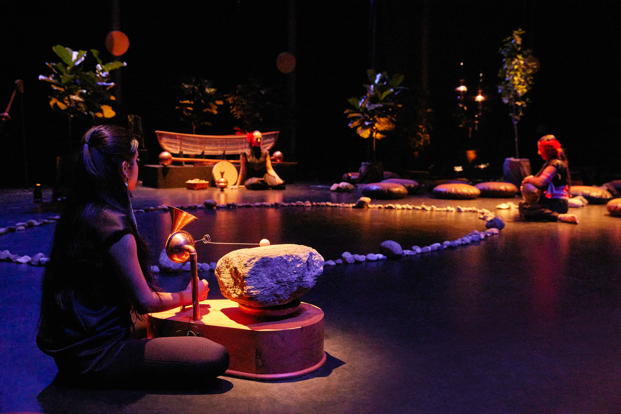 Two performers seated on a stage bathed in dark purple and orange light. On the floor is an instrument on a stand with a rock, a circle of pebbles, a boat and potted plants.