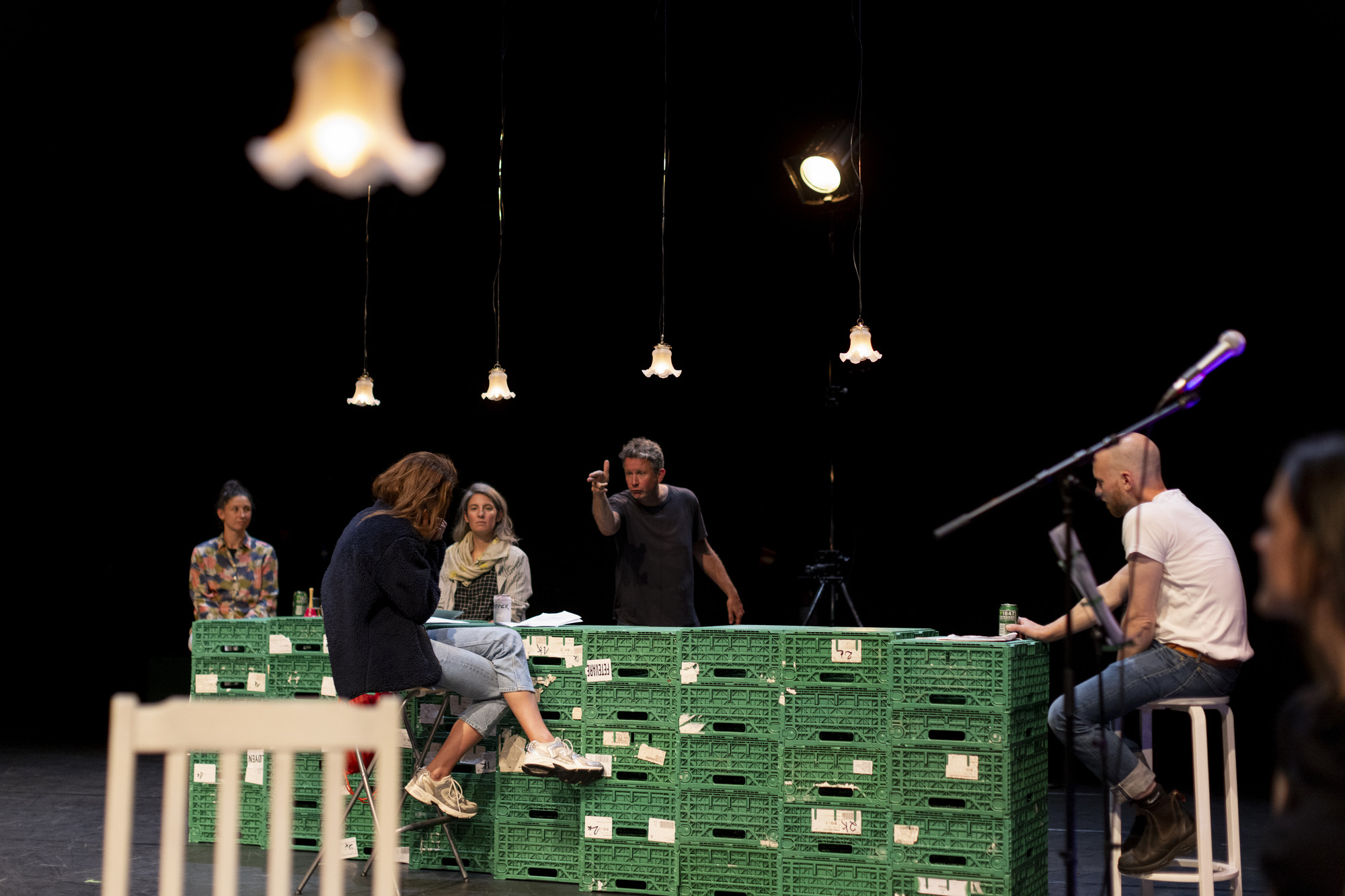 Performers rhearsing on a stage, sitting around a table made of green crates. One performer makes a hand gun gesture at another actor.