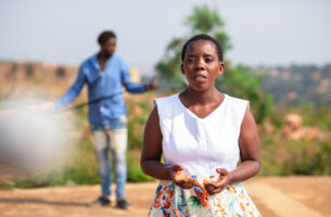 A Black performer stands outdoors, facing forward, caught in a moment of deep thought. Behind her, out of focus, stands a second performer against a backdrop of the dusty browns of dry dirt and the deep greens of the trees and shrubs that grow from it.