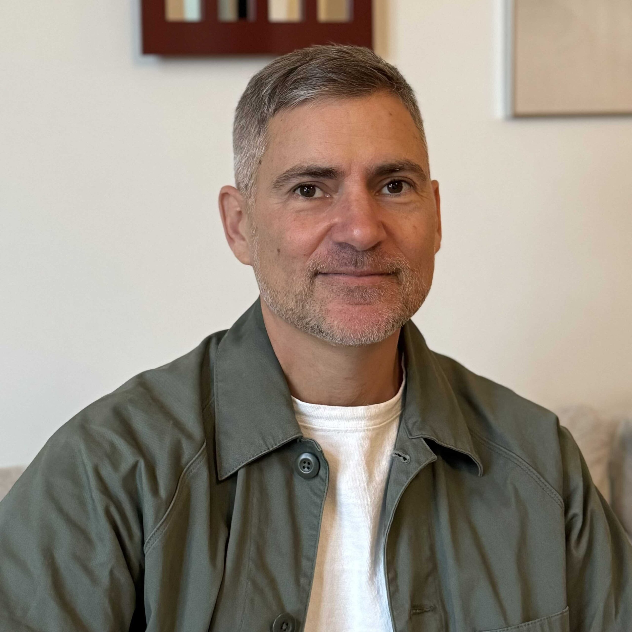 Diego Gil, a man with short grey hair and stubble, facing the camera and smiling sightly. He is wearing a gray-green jacket and white t-shirt.