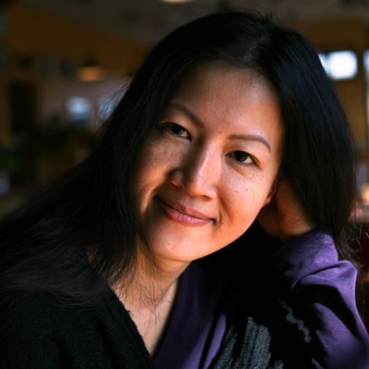 Janice Poon, a woman wearing a purple shirt, who is smiling and looking at the camera. Her head is slightly tilted and leaning on her hand. 