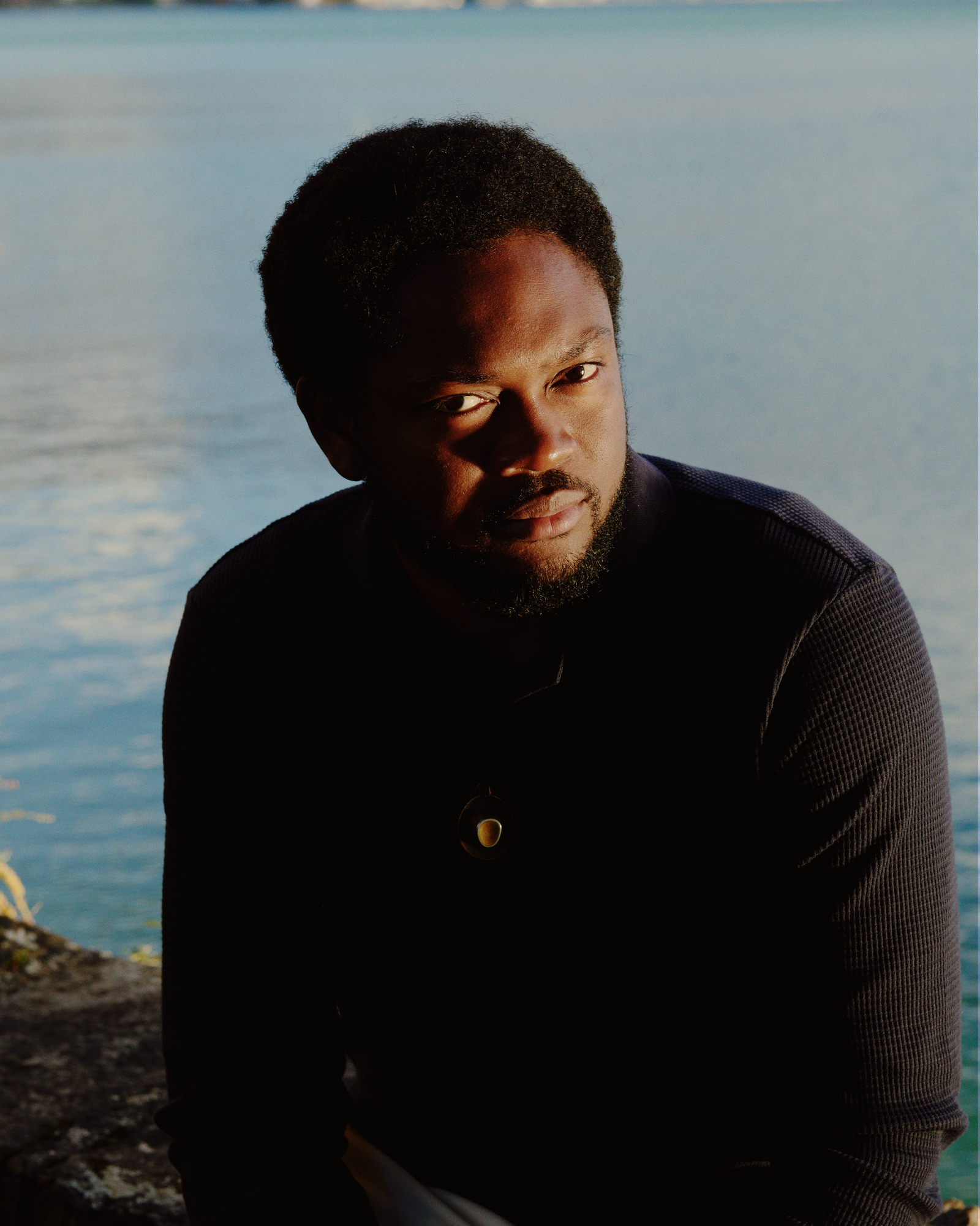 A Black man with short hair and beard, wearing a black sweater and necklace with shiny gem. Behind is a shimmering body of water.