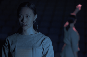 Two asian performers stand on an empty stage under unforgiving light. The first woman wears a severe, focussed expression, underscored by her futuristic structured grey top. In the background, the second performer, dressed in matching garb, faces away and reaches above her head.