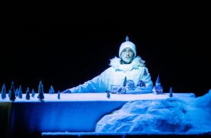 A man dressed for winter stares dead ahead with piercing eyes. He sits at a snow-white table scattered with miniature trees and a few tiny houses. His arm, which rests on the table, is an extension of the landscape, with little houses and trees sprouting from him as though he were a mountain.