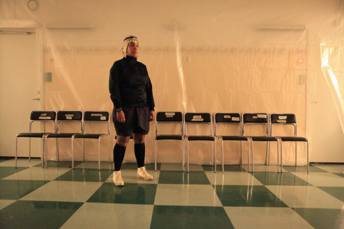 Wide shot of Majula Drammeh standing and looking to the right, wearing a metal headpiece and black sweatshirt and shorts with black knee socks and white sneakers. The room has a green checkered floor, a row of black and metal chairs and plastic sheeting against the wall. 