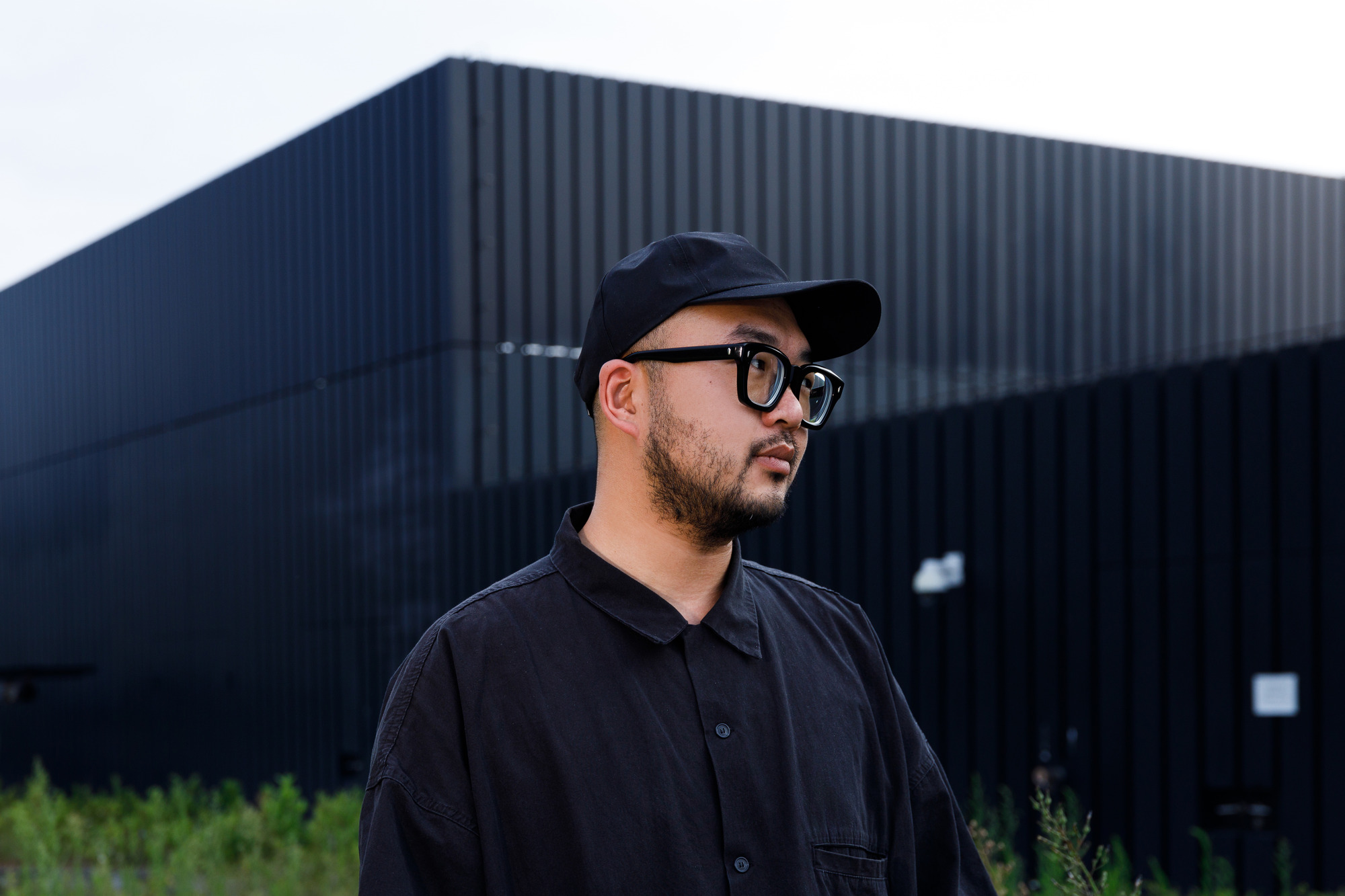 Wide shot portrait of River Lin looking off camera. He is wearing a black cap, chunky black glasses and a black button-down shirt and has a dark beard. In the background is a modern black building and grass peeking out below.