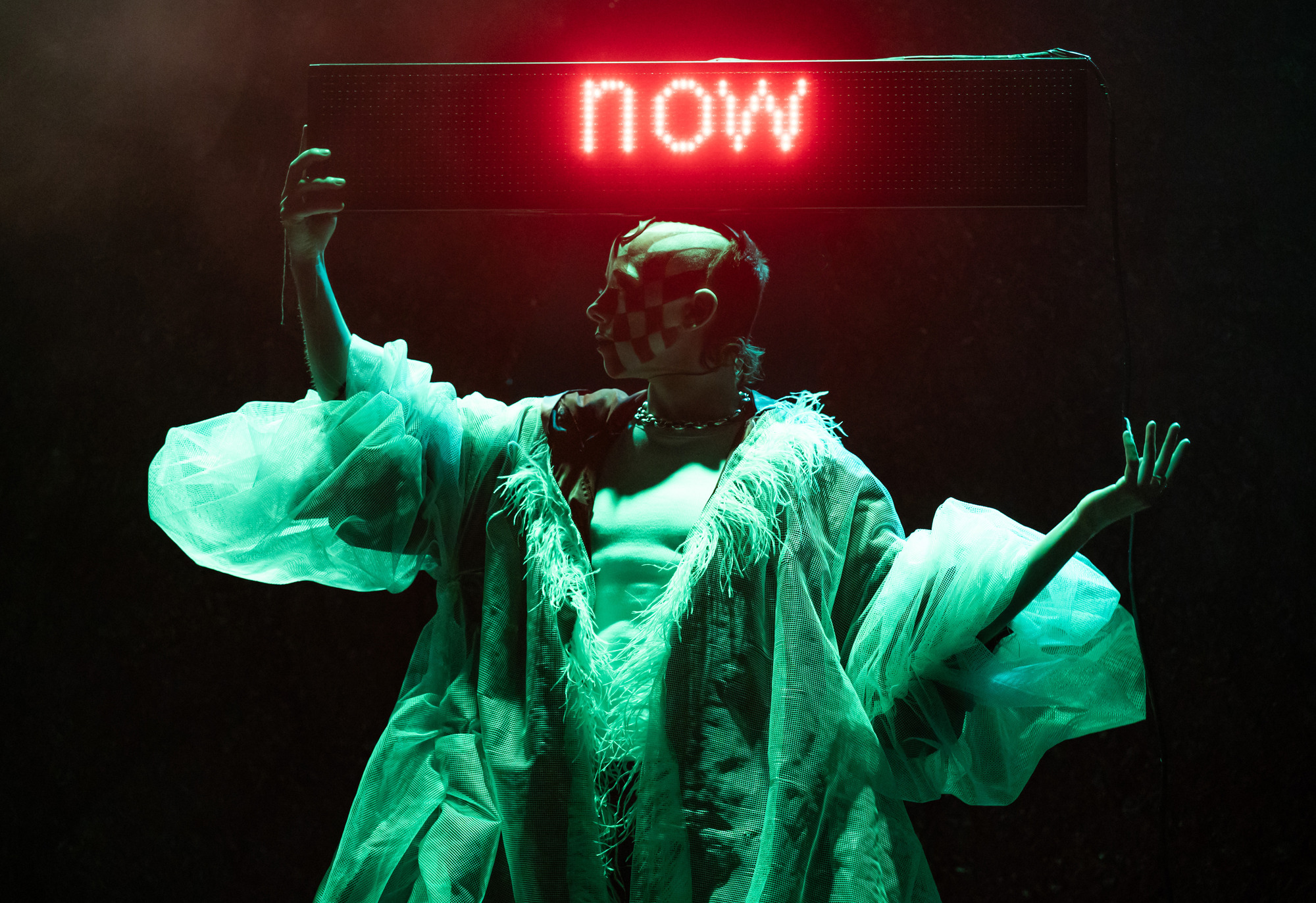 A performer bathed in green light, posing with hands raised. Their face is painted with a checkerboard pattern, and they are wearing a voluminous sheer coat with fringed collar. They are holding a neon light strip that says "now".