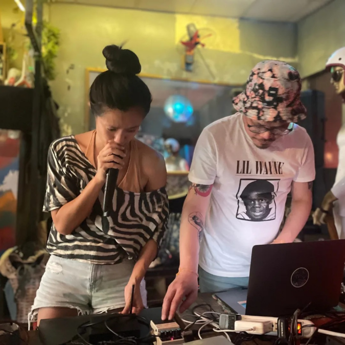 Two performers standing at a DJ deck, performing. On the left, Kim-Sanh-Chau has a black topknot and wears a zebra-patterned off-the-shoulder shirt and denim shorts, and is rapping into a microphone. On the right, Chittakone-Baccam is wearing glasses, a floral bucket hat and white Lil Wayne t-shirt and using a laptop  and music controller.