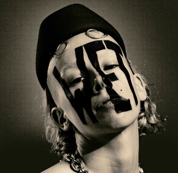 Sepia portrait of Wet Mess. They are looking at the camera with head tilted back, the word "WET" painted in black across their entire face. They wear a black cap and large chain necklace.