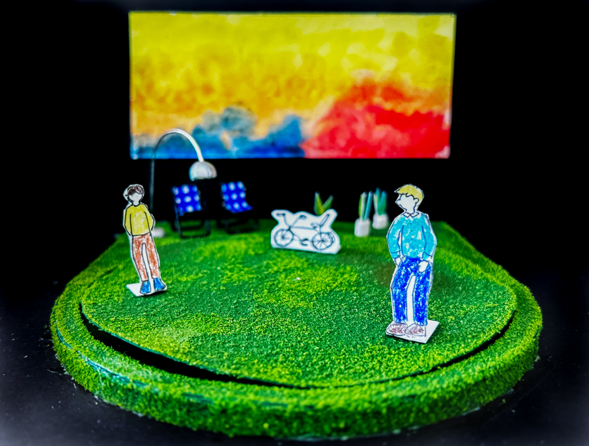 Miniature diorama of a round textured green pedestal with paper decorations: two human figures, a bike, and a pair of seats with a lamp. The yellow, blue and red background is out of focus.