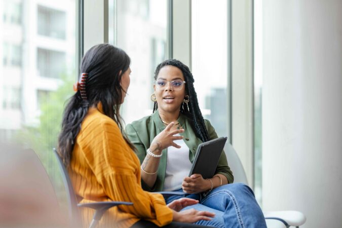 Two seated people talking against a glass window. On the left, a woman with long dark hair and a yellow shirt faces away, listening. On the right, a Black woman wearing glasses and a green jacket while  holding a tablet speaks to her with emphatically. 