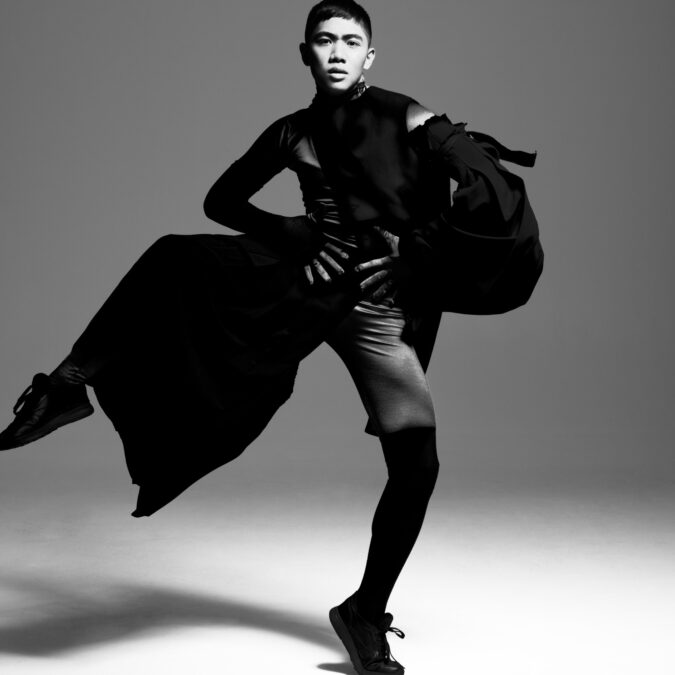 Black and white full body portrait of Ralph Escamillan dancing with one leg raised, wearing a flowing black garment